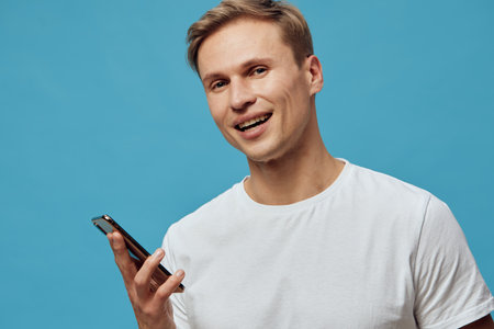 smiling young caucasian man hold smartphone use voice assistant gesture blue background casual white...