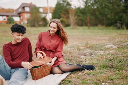 Candid outdoor picnic of a young couple sharing a wicker basket, conveying authenticity and credibility through genuine smiles, natural gestures and relaxed everyday connection in autumn.の写真素材