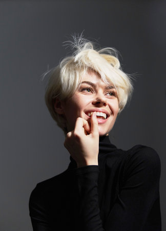 Smiling young woman with short blonde hair, wearing a black turtleneck, playfully biting her finger....