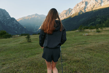 A solitary woman stands in a grassy alpine meadow, facing distant mountains as sunlight warms the scene, creating a peaceful outdoor moment, with quiet skies and expansive horizon.の写真素材
