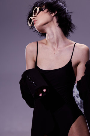 Stylish woman in black bodysuit posing with sunglasses and short hair on purple backgroundの写真素材