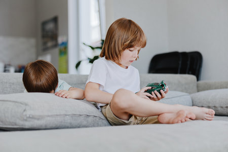 Engaging boy playing with a toy car while his younger sibling rests on the couch, surrounded by a cozy living room atmosphere filled with natural light.の写真素材