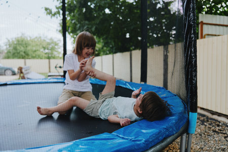 Playful children enjoying a trampoline, laughing joyfully in a sunny backyard, surrounded by greenery and a bright atmosphere, embracing fun and adventure.の写真素材
