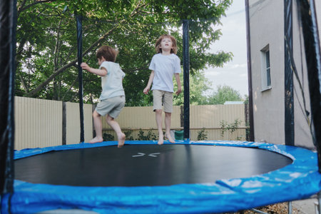 Two joyful children playing on a trampoline in a sunny backyard, capturing the essence of fun and carefree summer days with bright greenery around.の写真素材