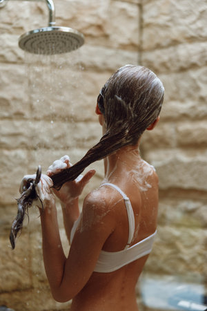 Woman washing long hair under outdoor shower with natural stone wall background, water droplets, soap suds, and relaxed summer vibe.の写真素材