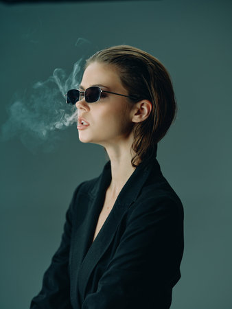 Young stylish woman wearing sunglasses smoking with smoke cloud on moody dark backgroundの写真素材