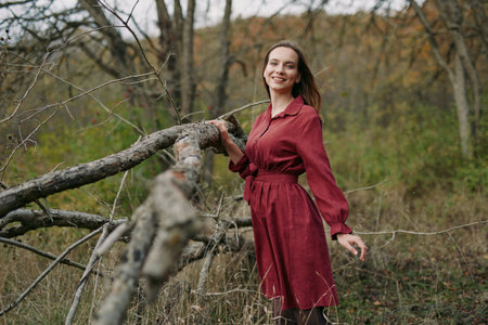A candid environmental portrait of an adult woman in a red dress standing by a fallen tree in woodland, conveying authenticity through a genuine relaxed smile and natural light mood.の写真素材