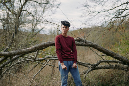 Authentic portrait of a young man standing in a rural landscape beside a fallen tree, natural light, casual maroon sweater and denim, conveying genuineness and credibility.の写真素材