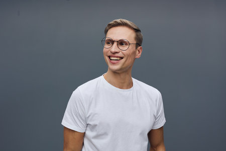 Smiling young man wearing glasses in casual white t-shirt looking away isolated on plain gray...