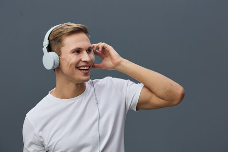 Smiling man wearing white t-shirt and wireless headphones, holding one hand near ear, enjoying...