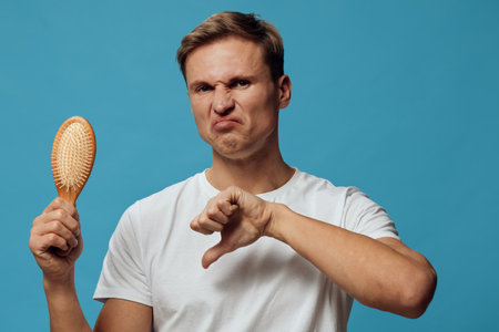Smiling man with a disgusted expression holding a hairbrush in one hand and showing a thumbs down gesture with the other, on blue background. Humorous casual portrait, negative facial expression, white t-shirt, indoor setting.の写真素材