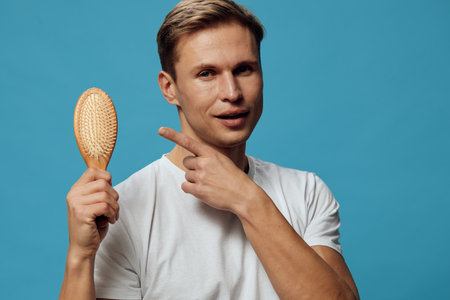 Male young adult with short brown hair holding a hairbrush and pointing at his cheek, casual white t-shirt, standing against a solid turquoise background, studio portrait, humorous grooming concept.の写真素材