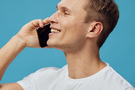 Smiling young man talking on black mobile phone isolated on blue background. Casual clothing, cheerful expression, close-up portrait capturing communication and technology lifestyle concept.の写真素材