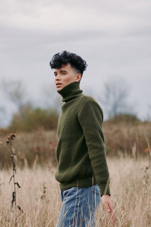Young man in a textured green sweater stands in a windswept field, conveying a natural, honest...