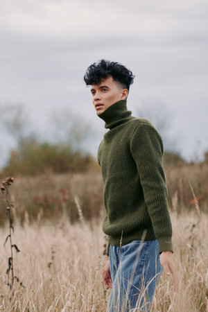 A young man stands in a grass field wearing a dark green knit turtleneck, exuding authenticity and quiet confidence in a natural portrait with soft light and minimalist backdrop.の写真素材