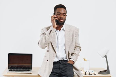 Professional young African American man in business casual attire talking on mobile phone while standing at office desk with laptop, glasses and writing tools, studio shot. Business communication conceptの写真素材