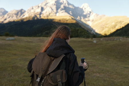 A lone traveler with a large backpack stands in a wide meadow as rugged mountains rise behind, creating a serene outdoor scene and a sense of exploration.の写真素材