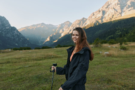 A cheerful woman hiker stands in a wide alpine meadow with mountains in the distance. She wears a...