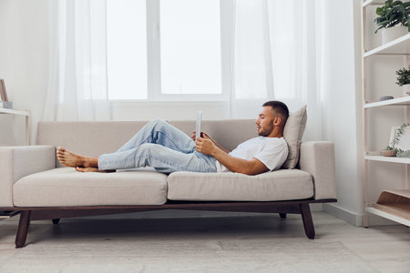 Relaxed man reading a book on a cozy sofa in a bright, modern living room. The natural light enhances the tranquil atmosphere and serene mood.の写真素材
