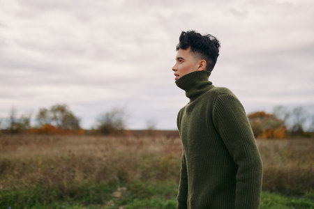 A genuine portrait of a young man in a thick olive sweater standing in an open field, side profile capturing natural expression and unposed stance, conveying authentic, realistic character and quietの写真素材