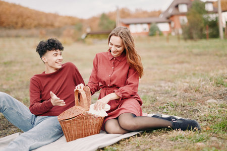 A candid outdoor picnic scene showing a young couple sharing bread from a wicker basket, genuine smiles and relaxed posture conveying authenticity and visual credibility in a rustic setting.の写真素材