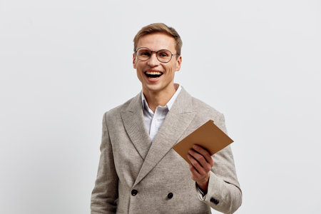 Smiling young man with glasses dressed in a beige blazer and white shirt holding a notepad, looking cheerful and confident, standing against plain white background. Studio portrait, people lifestyle concept.の写真素材