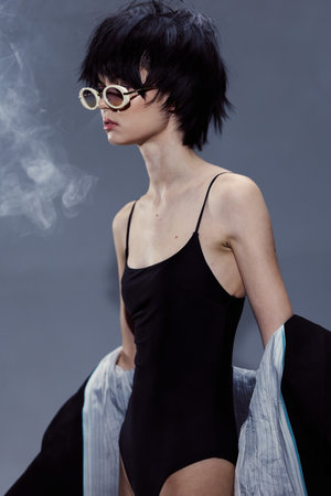 Slim woman wearing black swimsuit and sunglasses posing with jacket on gray background with smokeの写真素材