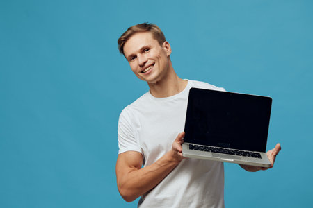 Smiling man holding laptop with black open screen, casual white t-shirt, young adult, light brown hair, cheerful emotion, isolated on blue background, studio portrait, technology concept.の写真素材