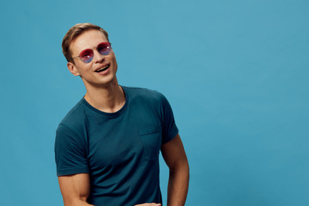 Smiling young caucasian man wear sunglasses teal t-shirt standing isolated on blue background casual...