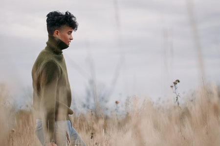 Young man in a textured sweater walking through tall dry grass, photographed in a candid, natural moment that emphasizes genuine presence and visual realism in soft overcast light.の写真素材