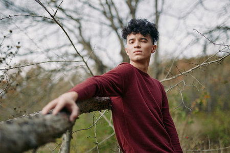 A candid portrait of a young man in a maroon sweater leaning on a wooden fence, radiating authenticity and quiet credibility in a calm natural setting.の写真素材