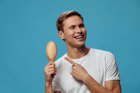 Smiling young caucasian man pointing at hairbrush wear white t-shirt isolated on blue background studio portrait mental health gadgets conceptの写真素材