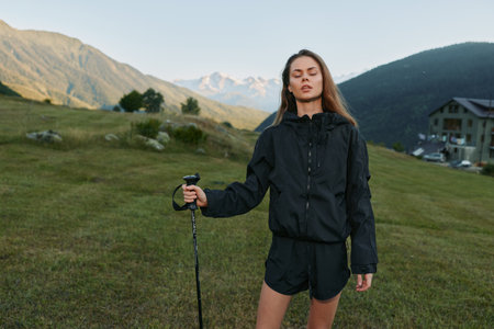 A confident young woman stands in a grassy field with a trekking pole, wearing a black windbreaker and shorts. She looks ahead, ready for a hike under clear skies.の写真素材