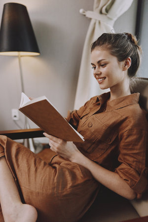 Young woman reading a book in a cozy brown dress, showcasing relaxation and happiness in a stylish...