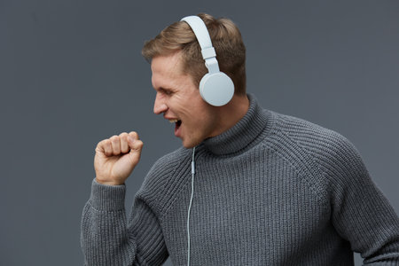 young caucasian man wearing gray sweater headphones singing enjoying music isolated on gray...