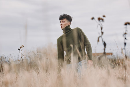 A candid portrait of a young man walking through tall dry grass in a green sweater, expressing quiet...