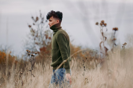 Young man walks through tall dry grass in a rustic meadow wearing a green turtleneck, captured in candid composition that highlights authenticity, credibility, natural texture and quiet, real emotion.の写真素材
