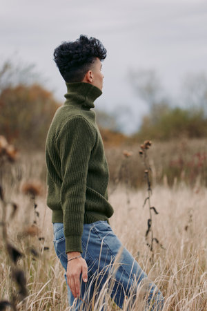 Young man in an olive knit sweater stands in a dried meadow, captured in an authentic, realistic...