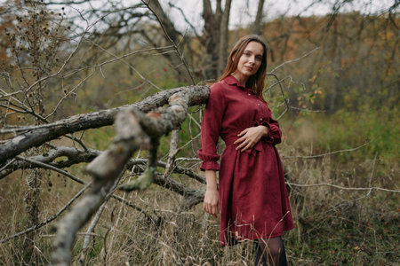 A candid portrait of a woman in a deep red dress leaning against a weathered branch, conveying authenticity and credibility in a quiet natural setting.の写真素材