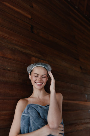 Smiling woman wrapped in towel and wearing shower cap stands happily against rustic wooden wall, enjoying spa moment with relaxed and joyful expression, cozy indoor setting.の写真素材