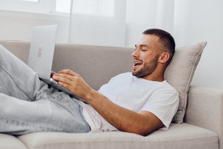 Smiling man working on a laptop, relaxed at home. Comfortable setting with a cozy sofa and natural...