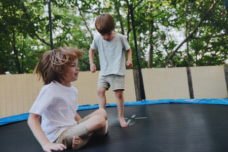 Two joyful boys play on a trampoline, experiencing energy and excitement in a bright outdoor setting surrounded by lush greenery and sunlight.の写真素材