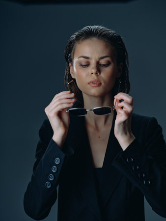 Young woman in black blazer holding sunglasses with wet hair, serious expression, studio portrait on dark backgroundの写真素材
