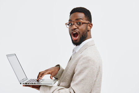 Professional young black man in casual blazer holding laptop and looking surprised or impressed, reacting to something on screen, isolated on plain background, studio portrait. Business technology conceptの写真素材