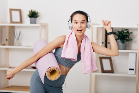 Young woman fitness enthusiast in activewear holds a rolled yoga mat and pink towel, wearing headphones and smartwatch, posing energetically in a bright home workout space.の写真素材