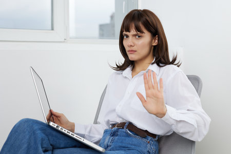 Woman sits with a laptop on a chair in a bright room and raises her hand to signal stop, with a calm...