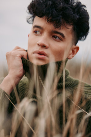 A candid portrait of a young man gazing away among tall grasses, wearing a chunky knit sweater. The image conveys genuine emotion, quiet focus, and authentic presence.の写真素材