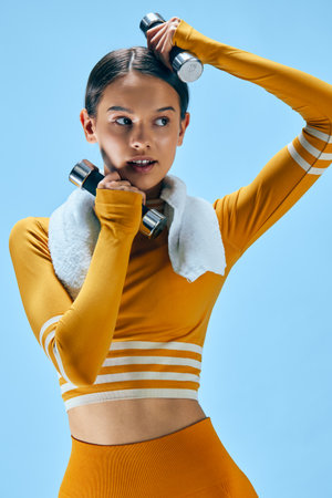 Fit young woman in yellow crop top and leggings holds chrome dumbbells with a white towel over her shoulders, performing focused arm workout against a blue studio background.の写真素材