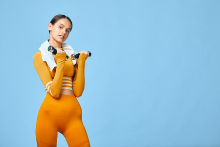 Young woman in orange activewear holds dumbbells with a white towel around her neck, posing...