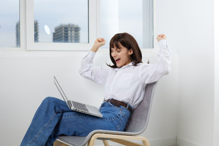Young woman sits in a modern chair by a window with a laptop on her lap, arms raised in celebration, conveying success and focus in a bright home office scene.の写真素材